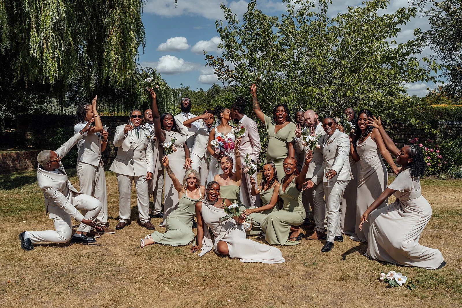 Kasey and Larisha kissing in the centre of their cheering wedding party during a fun group photo at Oaks Farm Weddings in Surrey.