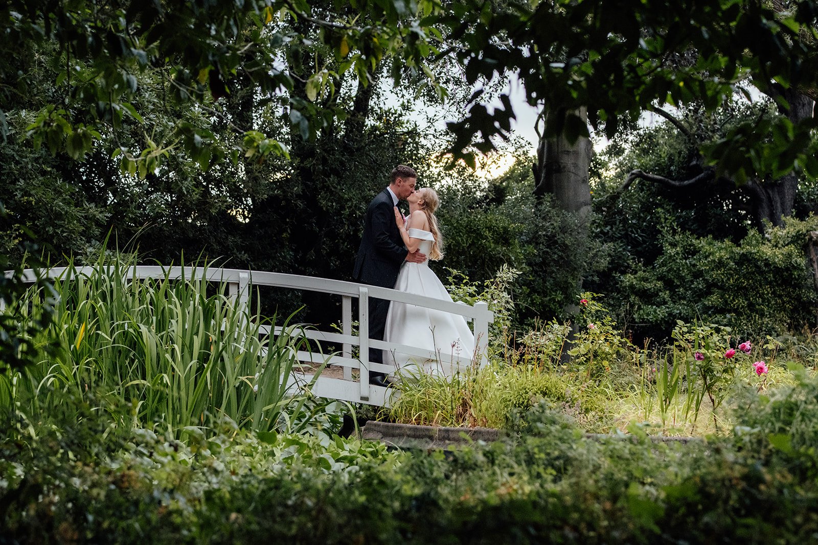Caitlin and Kevin sharing a kiss on a white garden bridge surrounded by greenery at Clevedon Hall in Somerset, captured during their evening wedding portraits.