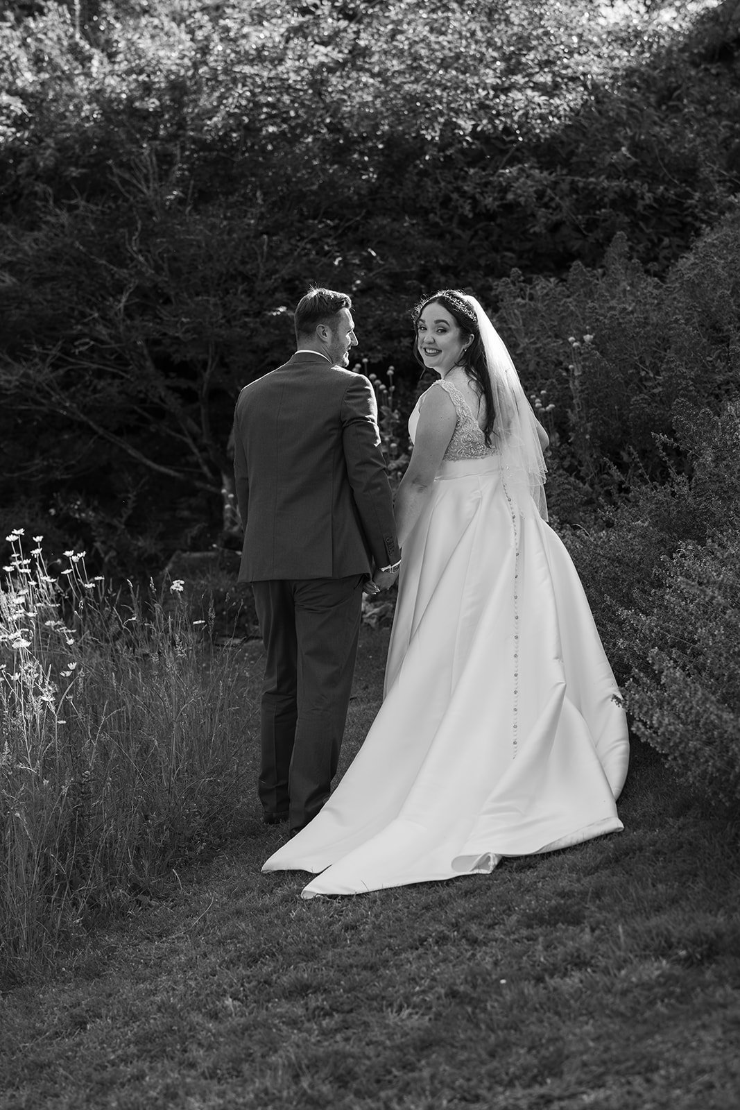 Black and white portrait of Chloe & Armani walking together at The Barn at Upcote in the Cotswolds, with the bride looking back towards the camera, photographed by Just James Weddings.
