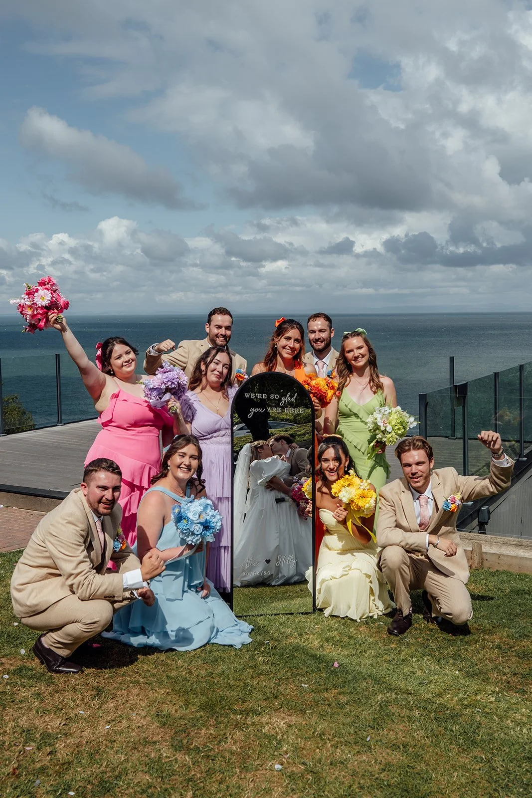 Holly & Alex’s wedding party pose around a personalised mirror with colourful bouquets and the North Devon coast behind them at Sandy Cove Hotel, photographed by Just James Weddings.