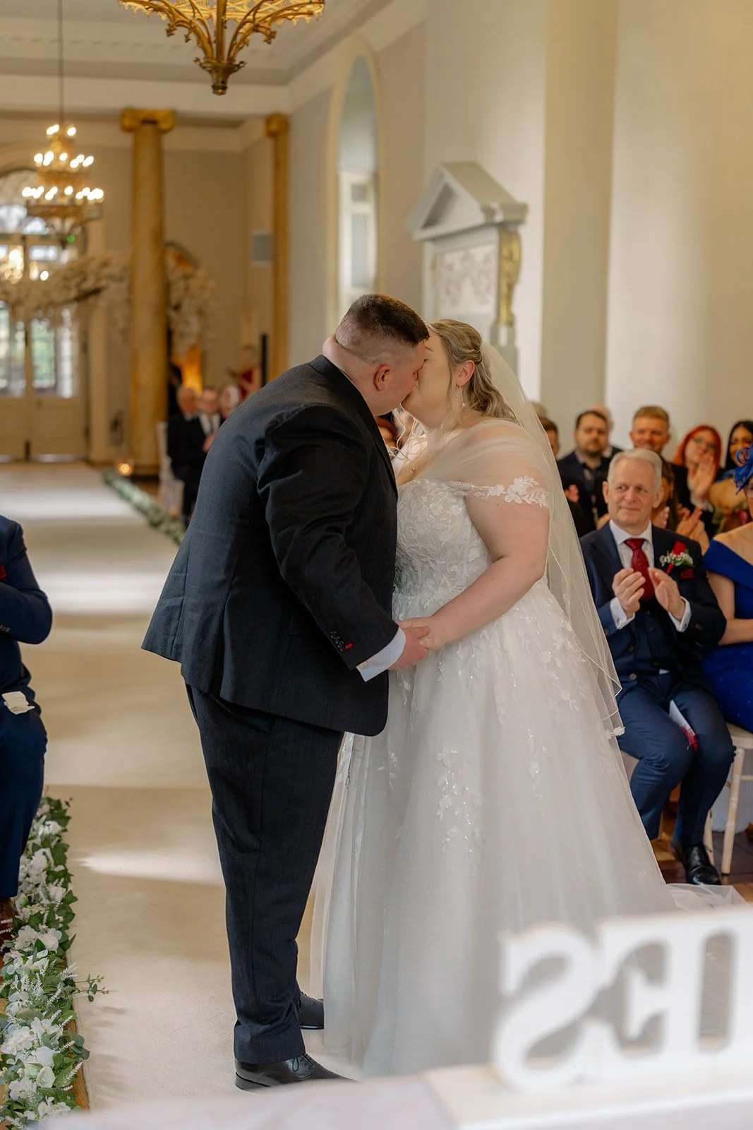 Becky & Rob share their first kiss during the ceremony at Clearwell Castle in Gloucestershire, UK while guests applaud from the aisle. Emotional moment captured during their Clearwell Castle wedding ceremony in the grand hall.