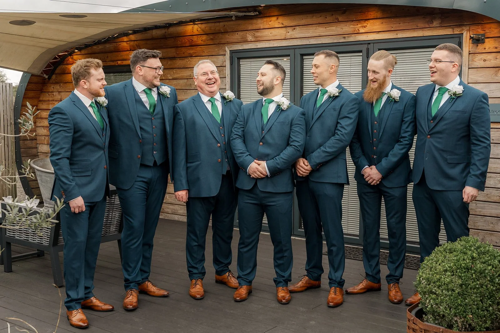 Groom with groomsmen and family laughing outside Kingscote Barn in the Cotswolds, UK, during Siobhan & Nicholas' wedding morning, wearing blue suits with green ties for relaxed rustic barn wedding photography before the ceremony on the terrace.