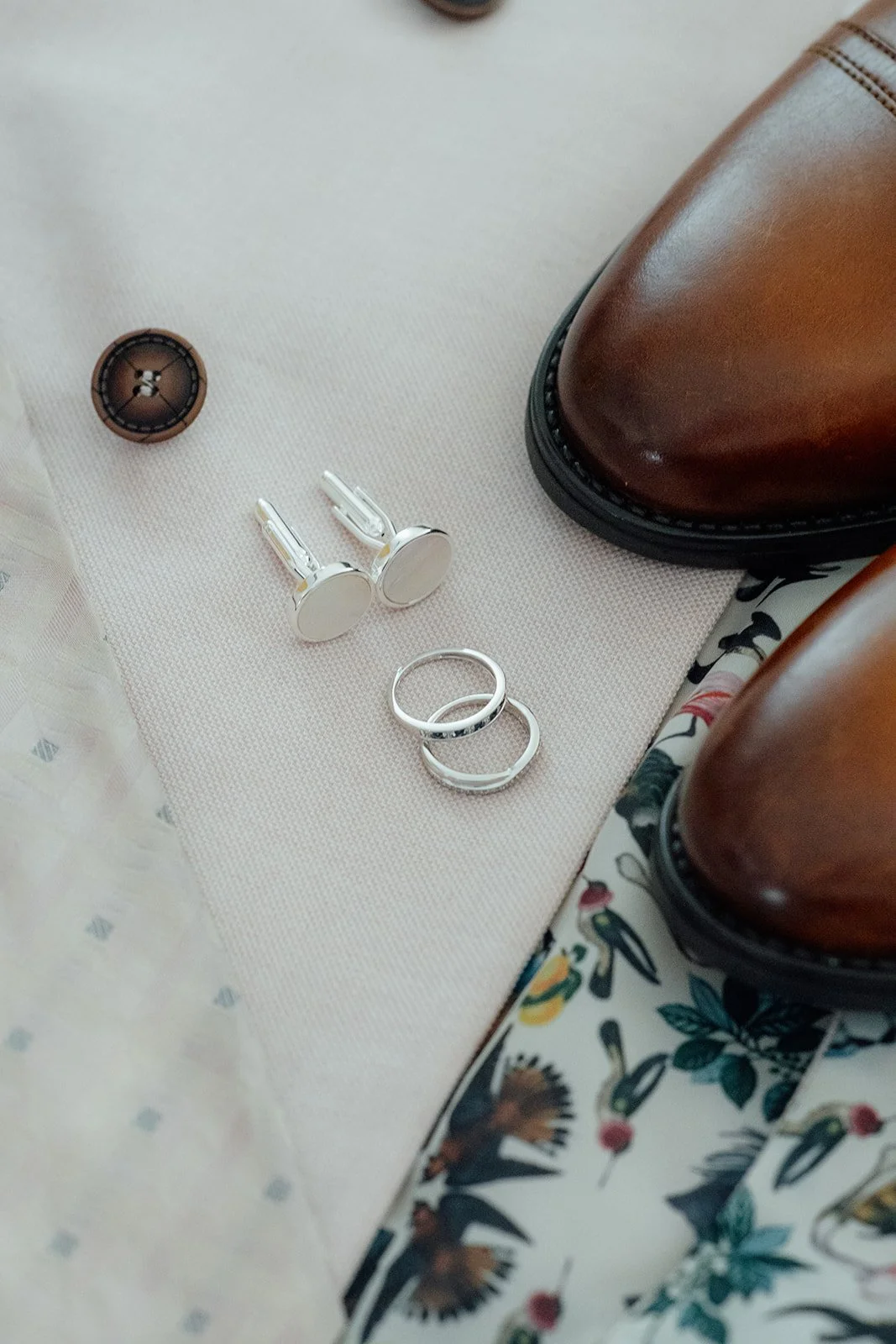 Kasey and Larisha’s wedding rings and silver cufflinks styled beside brown leather shoes and a floral tie at Oaks Farm Weddings in Surrey.