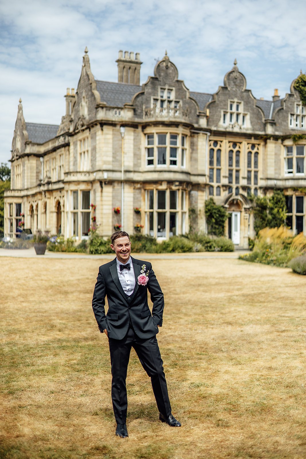 Kevin smiling in a black tuxedo with pink boutonnière on the lawn at Clevedon Hall in Somerset, with the historic wedding venue forming a grand backdrop.