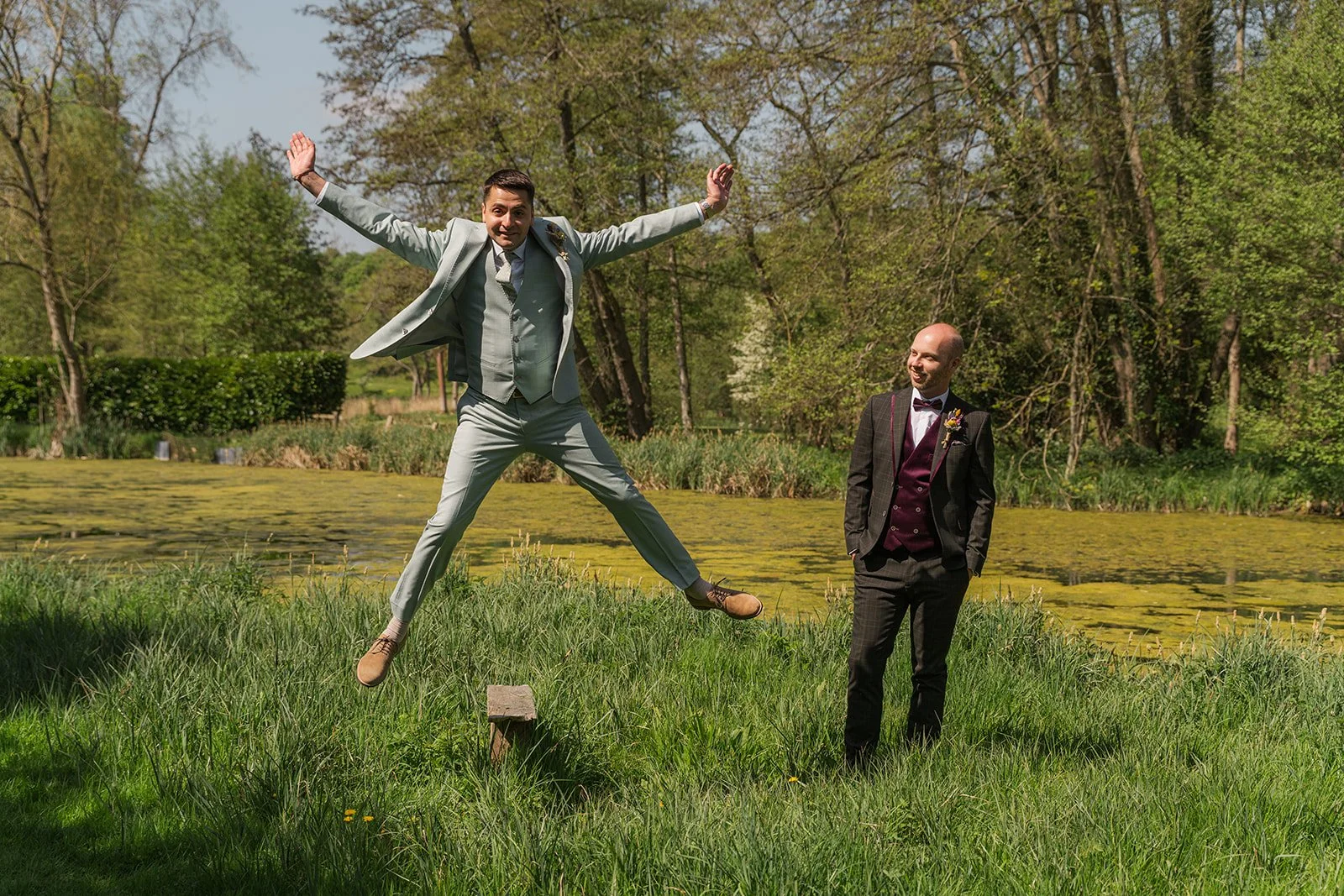 Gavin leaps into the air with his arms outstretched while Sam watches and laughs beside him on the grass at The Great Barn. The playful moment captures their joy and energy during relaxed outdoor wedding portraits surrounded by greenery.