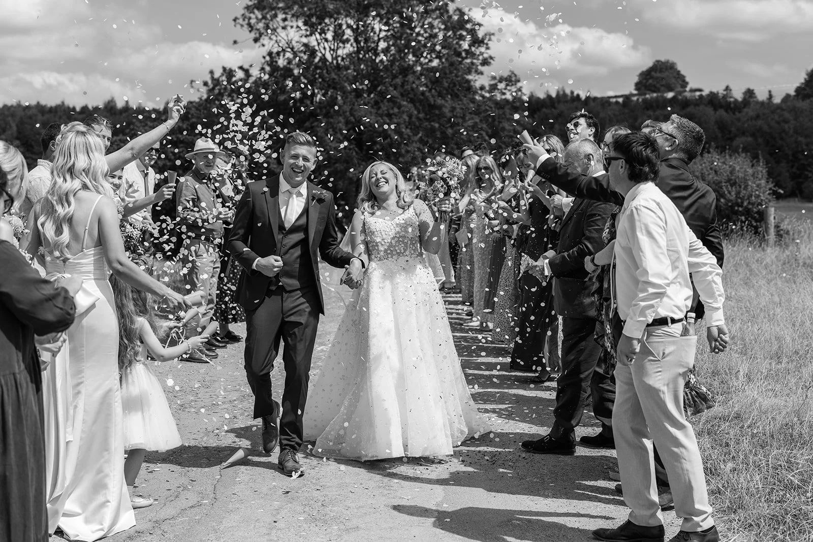 Black and white photo of Elise & Tom walking through falling confetti after their wedding at Bowerchalke Barn in Wiltshire, surrounded by cheering guests, photographed by Just James Weddings.