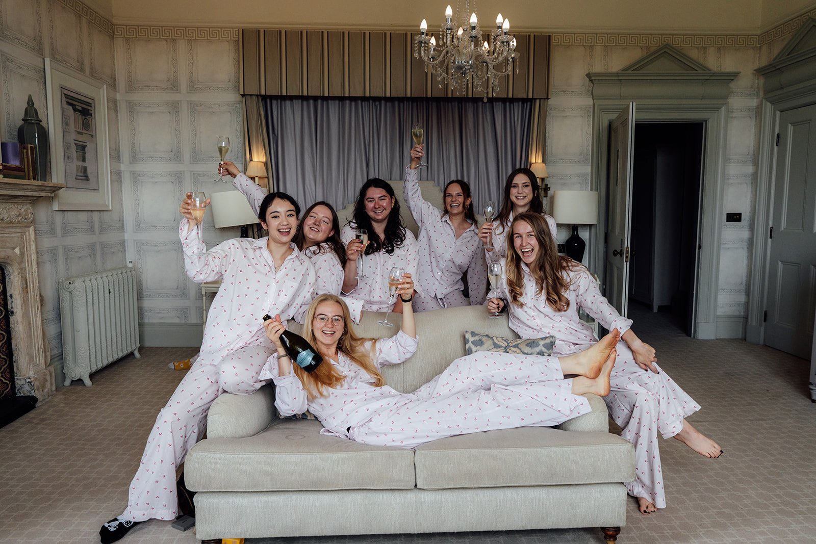 Caitlin and her bridesmaids celebrating before the wedding at Clevedon Hall in Somerset, wearing matching heart-print pyjamas while raising glasses of champagne in a bright bridal suite.