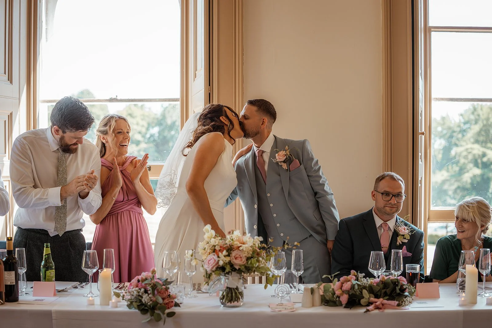 Michelle and Andy share a kiss at the top table during their wedding reception at Crowcombe Court in Somerset, UK, surrounded by floral arrangements and celebrating guests.