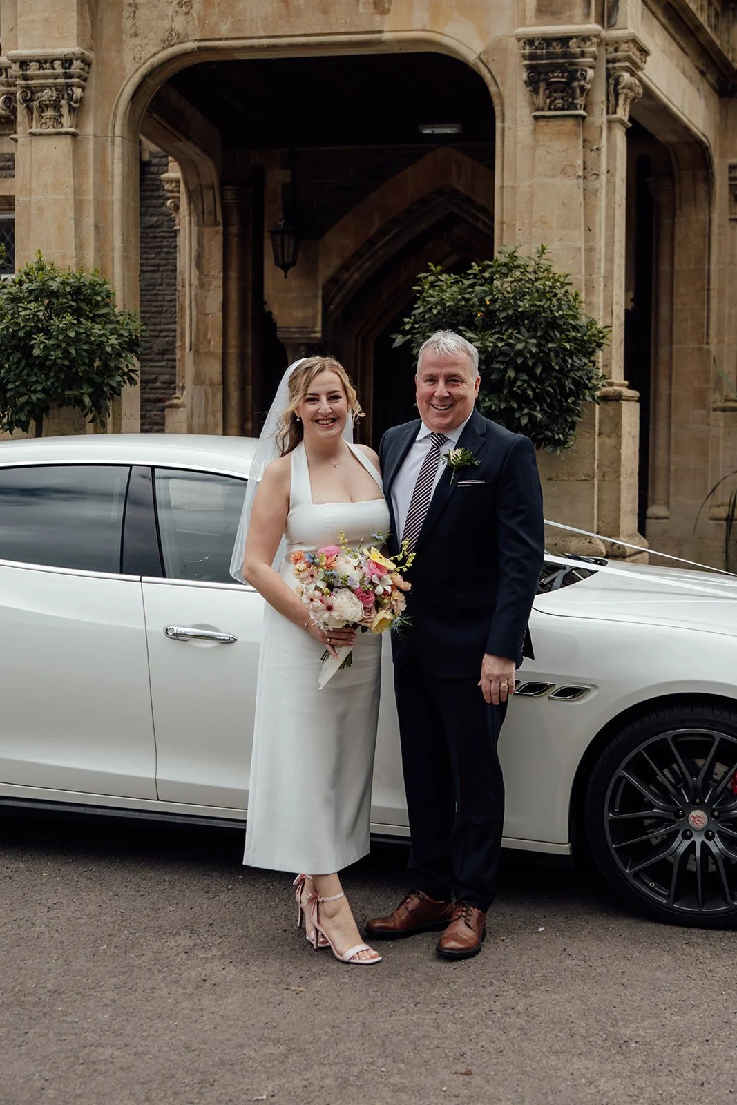 Bryony & Dad smiling outside Insole Court in Cardiff beside a white wedding car, with Bryony holding a colourful bouquet and the historic stone entrance creating an elegant backdrop for their relaxed wedding portraits together after arriving.