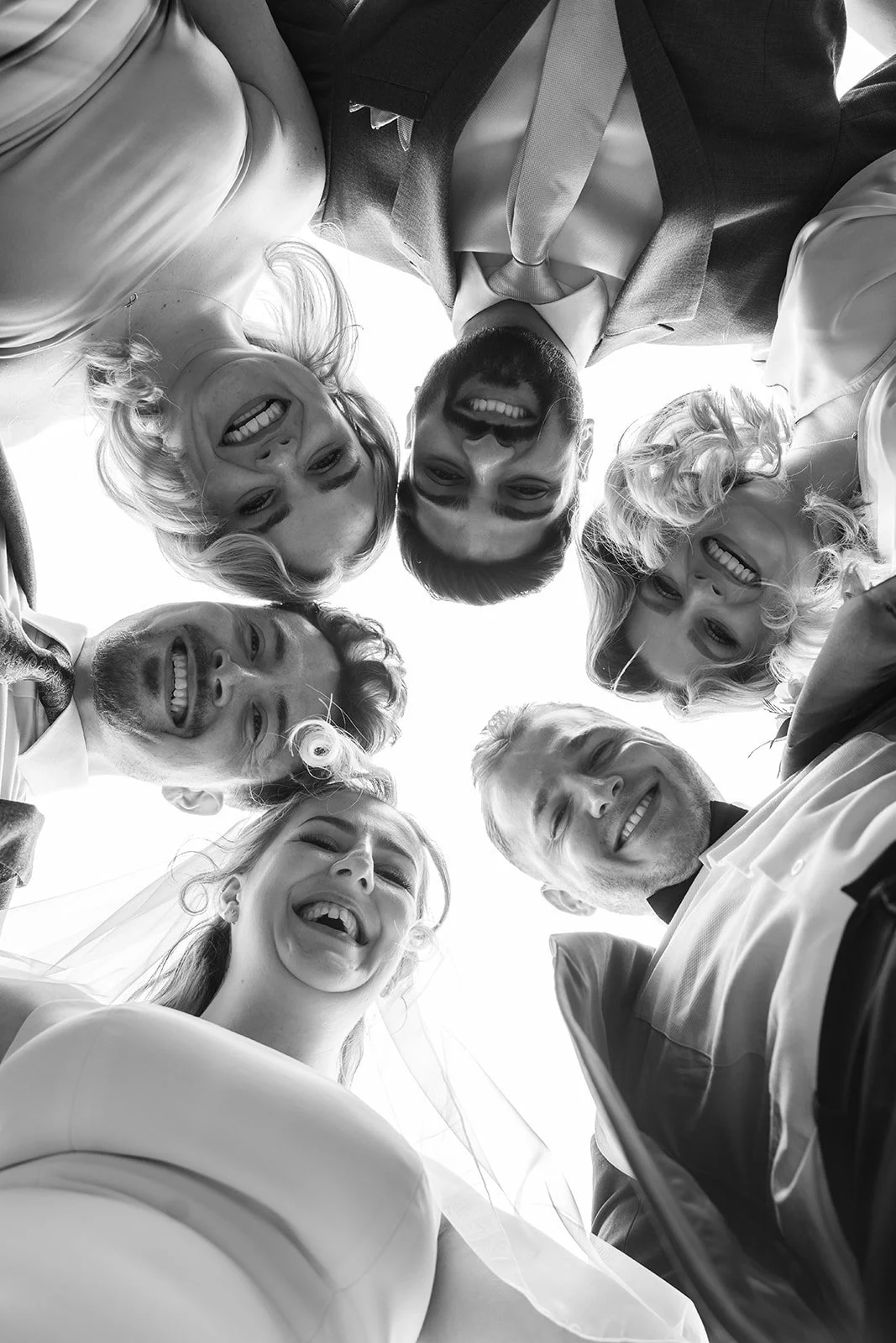 Bryony & Max laugh with close friends in a black and white low-angle group portrait at Insole Court in Cardiff, with smiling faces gathered in a circle above the camera for a playful and candid wedding party photo full of fun and personality.