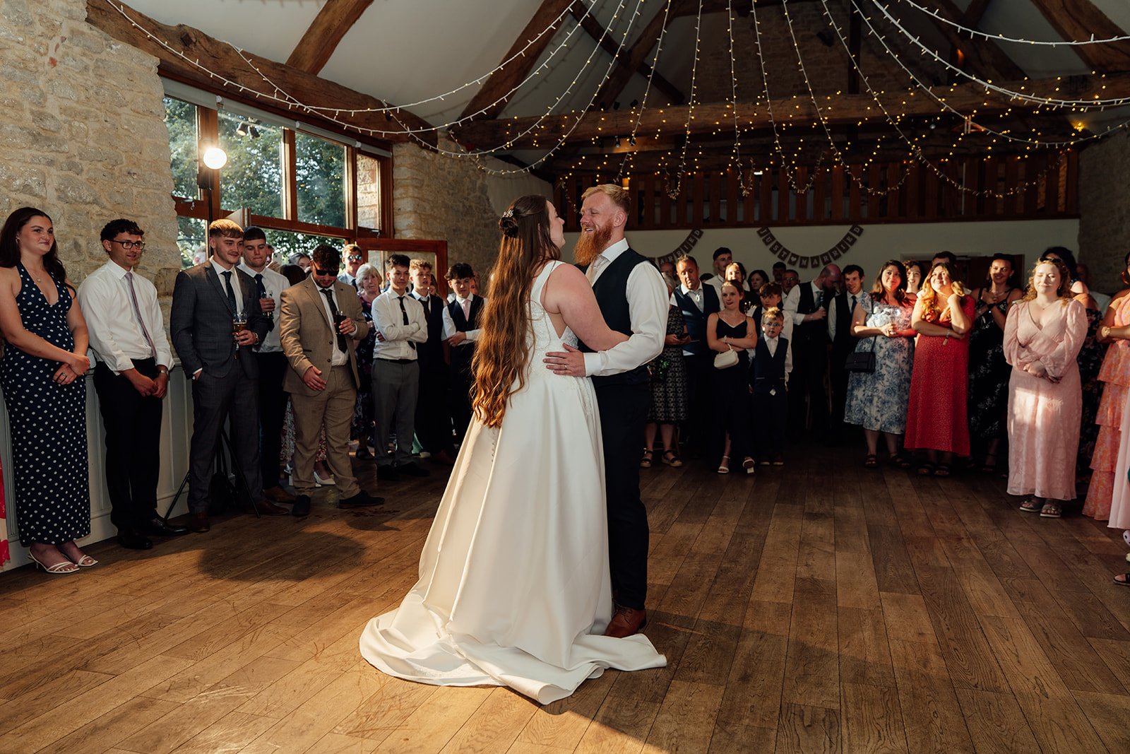 First dance for Louise & Jon at Wick Farm Bath in Somerset, UK, inside the rustic barn with wooden floors, stone walls, fairy lights, guests gathered around, warm evening sunlight, and a joyful newlywed moment on the dance floor together inside.