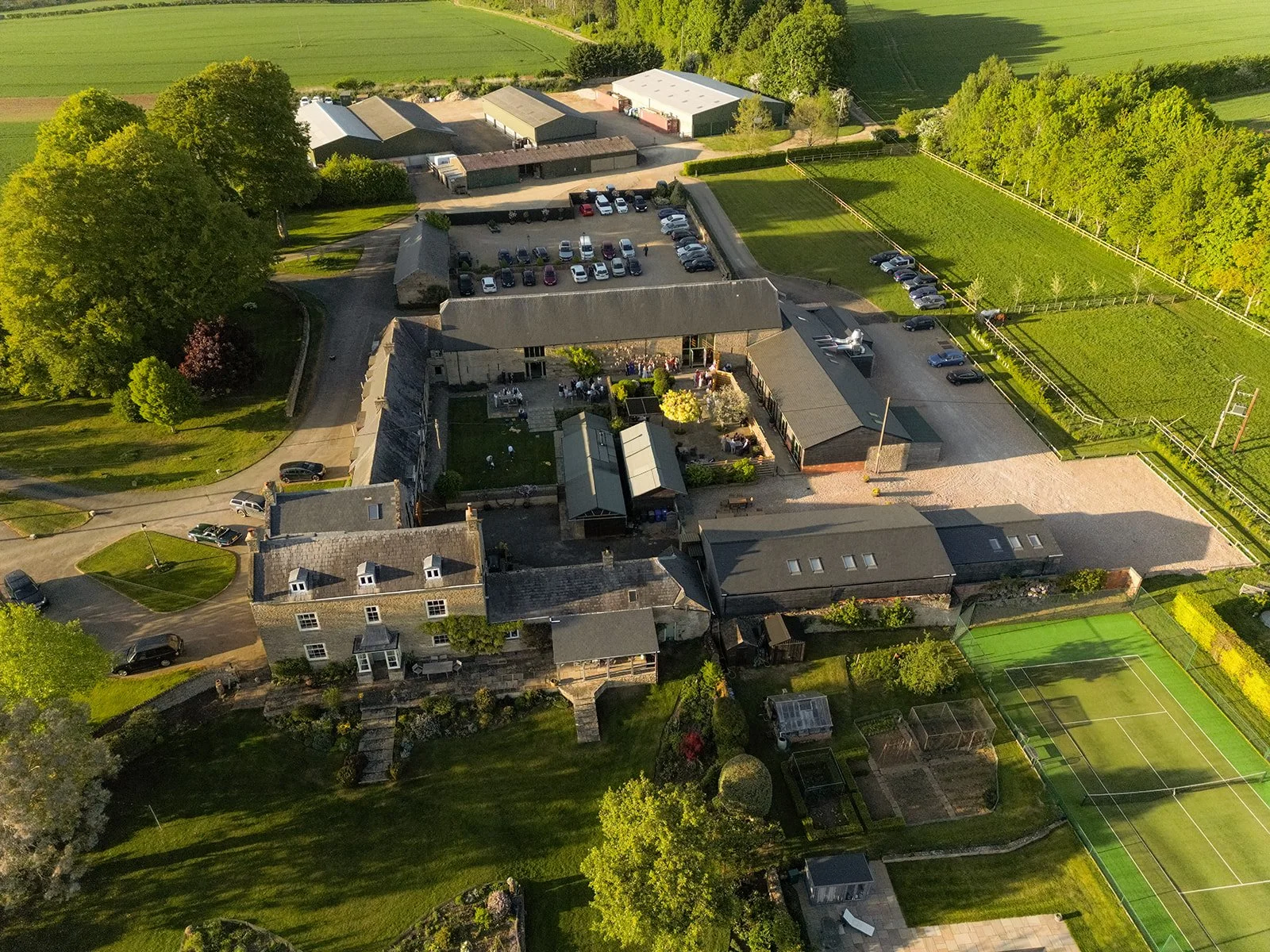 Aerial view of The Great Barn at Aynho in the Cotswolds, showing the stone barn, courtyard, parking, tennis court and surrounding countryside, photographed by Just James Weddings.