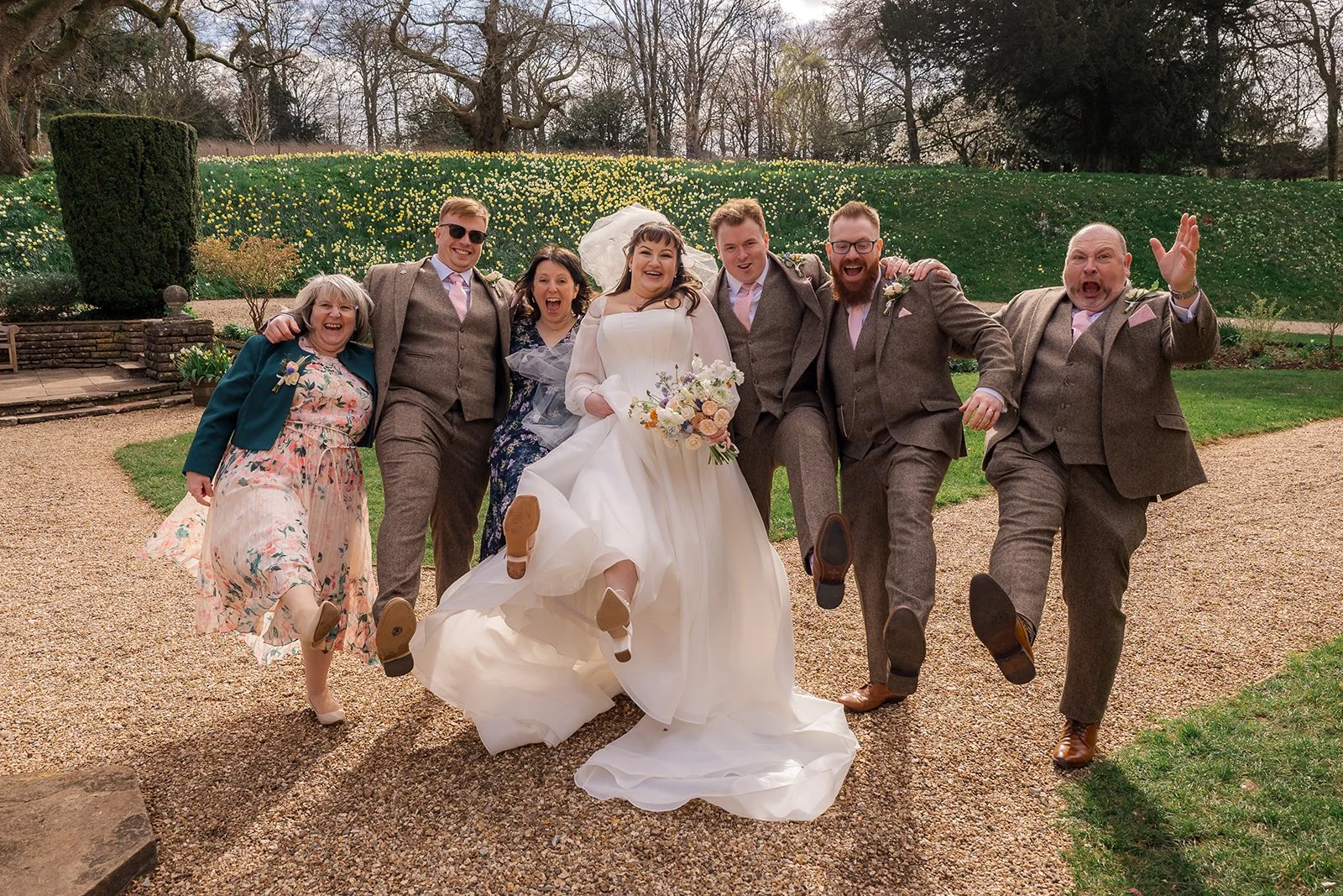 Emily & Josh pose with family and friends on the gravel paths at Coombe Lodge Blagdon, Somerset, UK, all lifting one leg and laughing together. Fun outdoor Somerset wedding group photo surrounded by gardens, daffodils and bright spring sunshine.