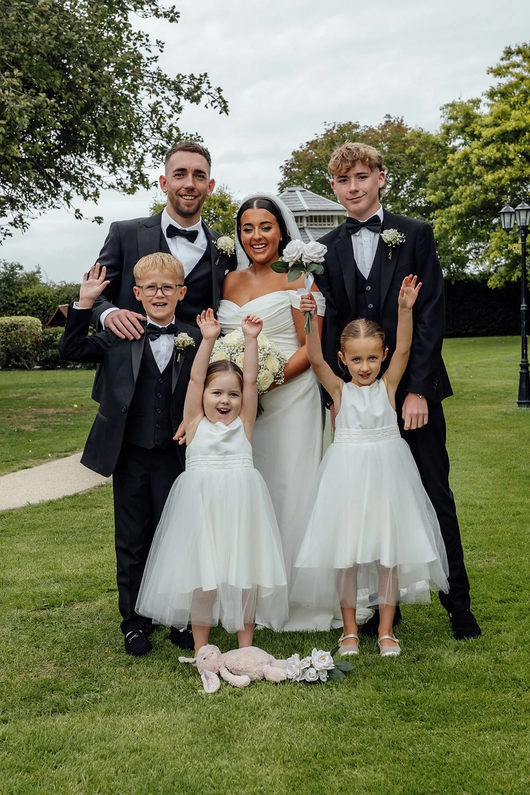 Nisha and Jack pose with four children from their wedding party on the lawn at Batch Country House in Somerset, UK, with the flower girls raising their hands while Nisha holds her white bouquet beside Jack, photographed by Just James Weddings.
