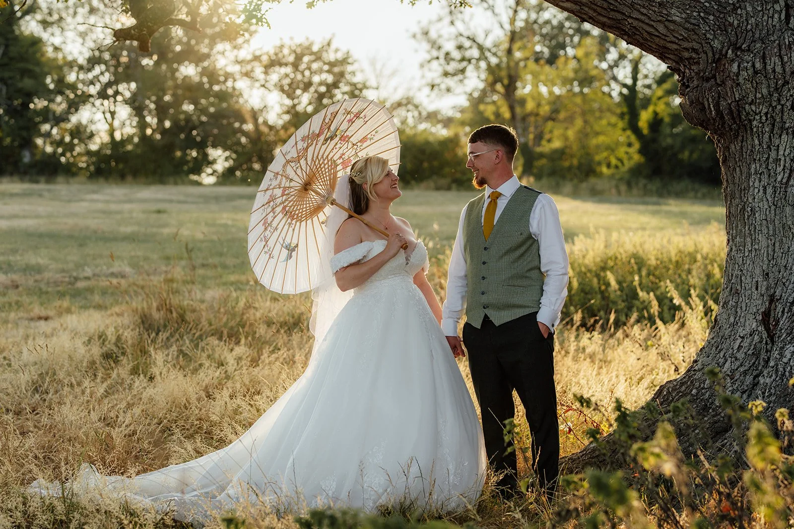 Lucy & Matthew share a relaxed golden-hour portrait beneath a large tree at Noah’s Pond in Somerset, with Lucy holding a decorative parasol, photographed by Just James Weddings.