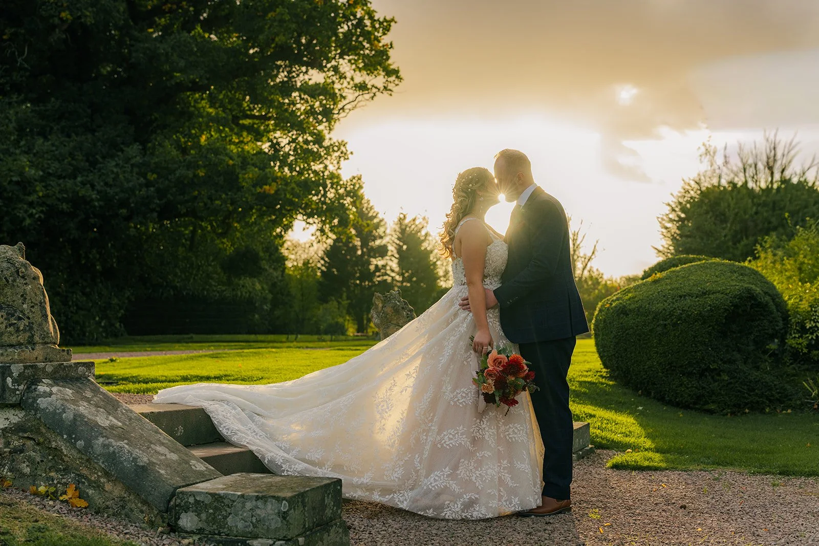 Lucy & Jay sharing a sunset kiss on the steps at Eastwood Park, gloucestershire. The bride’s lace gown glows in backlight while the groom holds her waist, with warm golden light, green lawns, and trees creating a romantic, peaceful wedding portrait.