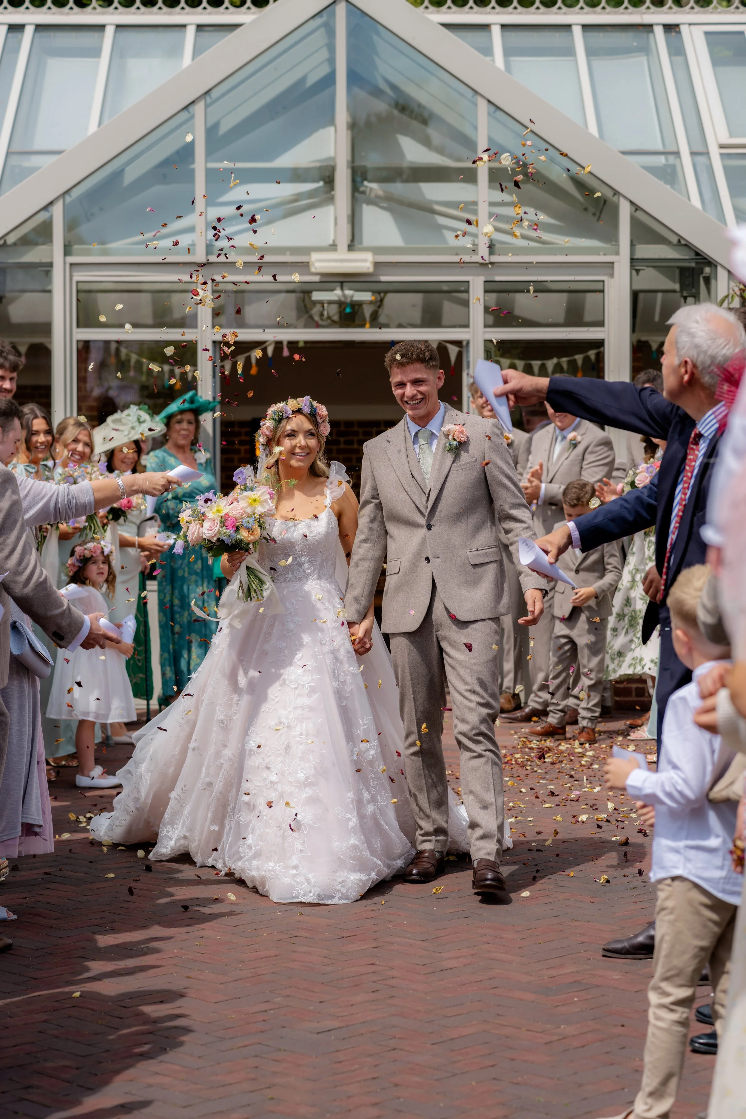 Jaye & Josh’s wedding at Syrencot in Wiltshire, captured as the newlyweds walk through a joyful confetti exit outside the glass orangery, surrounded by smiling guests, bright summer light and colourful petal celebrations after the ceremony.