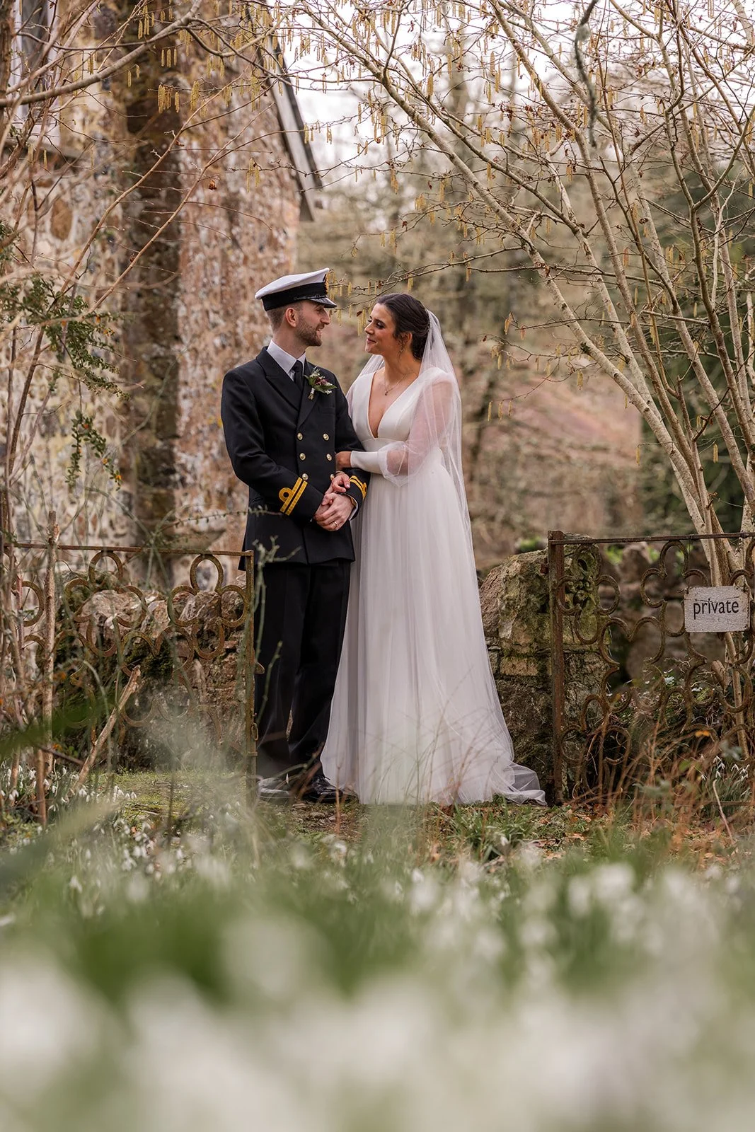 Megan & Luca stand beside an old stone wall and iron gate at The Great Barn Devon, framed by early spring branches and wildflowers. Megan rests her arm on Luca’s as they share a quiet look in this romantic Devon portrait.
