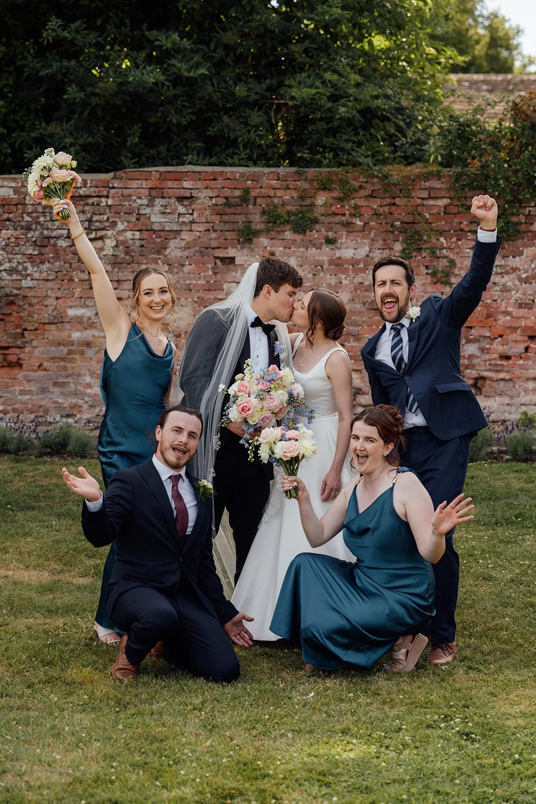 Rachel & Tom kiss as their wedding party cheer and raise bouquets around them in the gardens at Brympton House in Somerset, photographed by Just James Weddings.