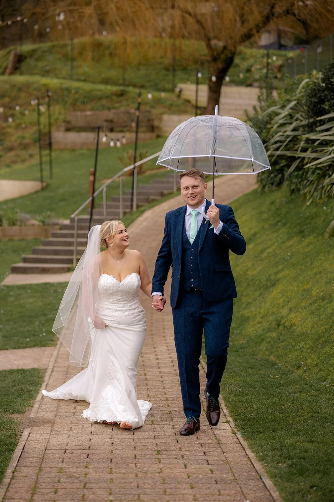 Andrew & Daisy walking hand in hand under a clear umbrella at Sandy Cove Hotel in Devon, smiling during light rain on the garden pathway, capturing romantic coastal wedding portraits at this clifftop seaside venue.