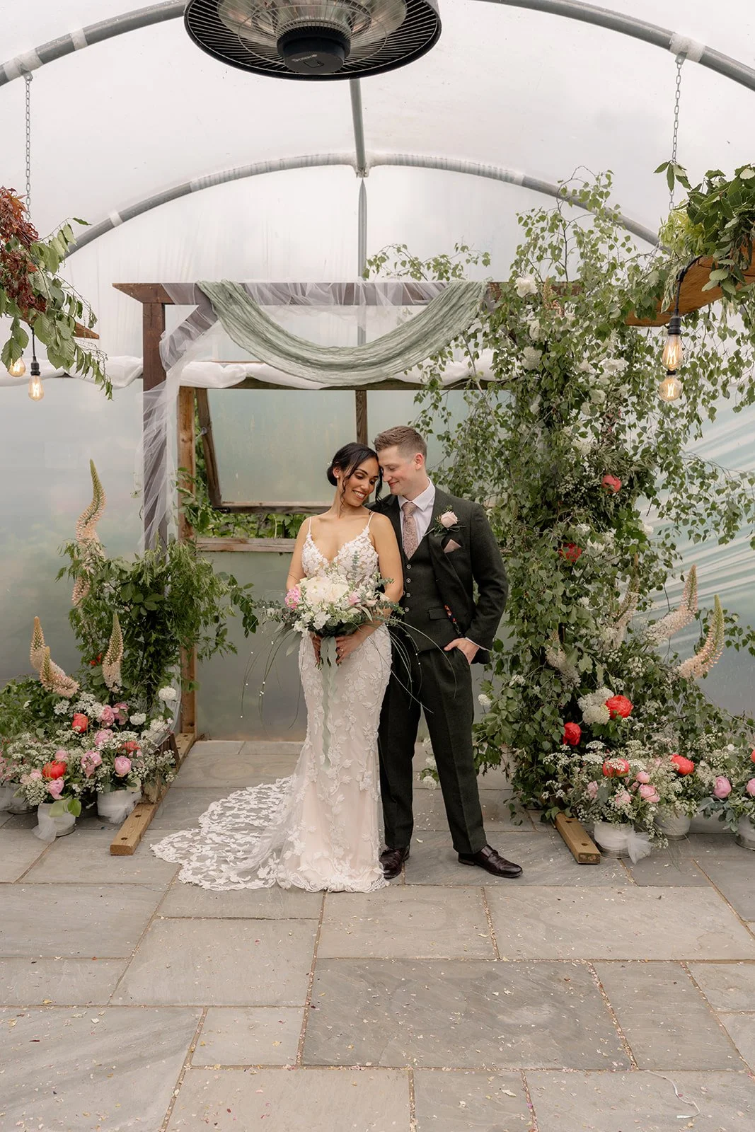 Jaye & Josh share a quiet couple portrait at Farrington's Farm in Bristol, standing beneath soft draped fabric, hanging greenery and warm festoon bulbs, with pink flowers framing their relaxed poly tunnel wedding ceremony backdrop beautifully.