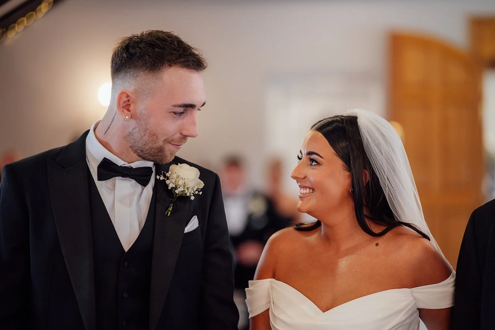 Nisha and Jack smile at one another during their wedding day at Batch Country House in Somerset, UK, with Jack in a black tuxedo and Nisha in an off-the-shoulder gown and veil during an indoor portrait, all photographed by Just James Weddings.