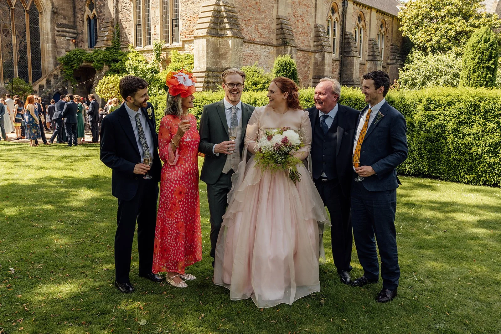 Charlotte & Jack family portrait at Bishop’s Palace in Somerset, UK, with the couple smiling on the lawn beside relatives in colourful wedding outfits, drinks in hand and the historic palace buildings and gardens behind them on a sunny day.