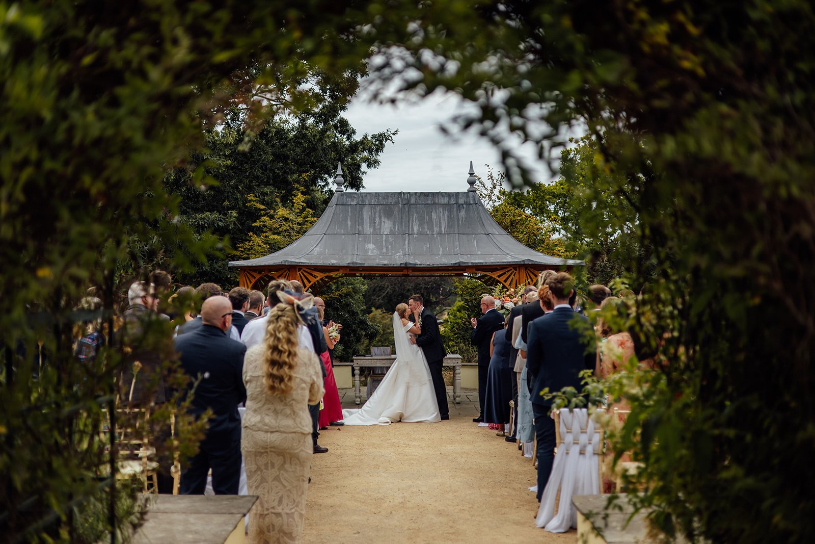 Outdoor wedding ceremony at Clevedon Hall in Somerset, with Caitlin and Kevin sharing a kiss beneath the garden gazebo as guests watch from either side of the aisle.