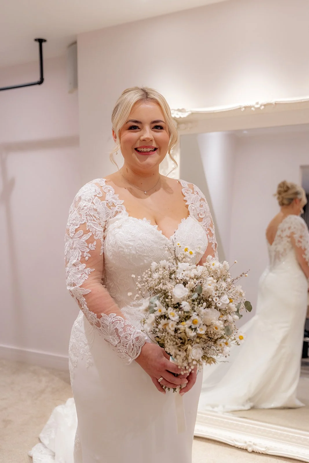 Daisy smiling with her bridal bouquet in her lace long-sleeve wedding dress at Andrew & Daisy’s Sandy Cove Hotel wedding in Devon, captured during morning preparations with mirror reflection highlighting her gown and natural bridal glow.