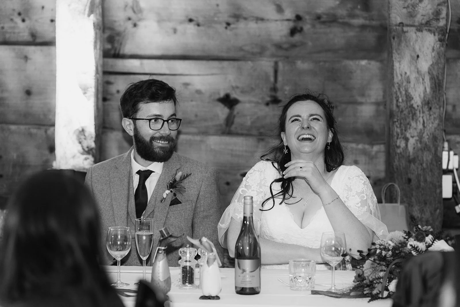 Black and white photo of Maisie & Andrew laughing during their wedding breakfast at Stockbridge Farm Barn in Dorset, photographed by Just James Weddings for candid reception moments.