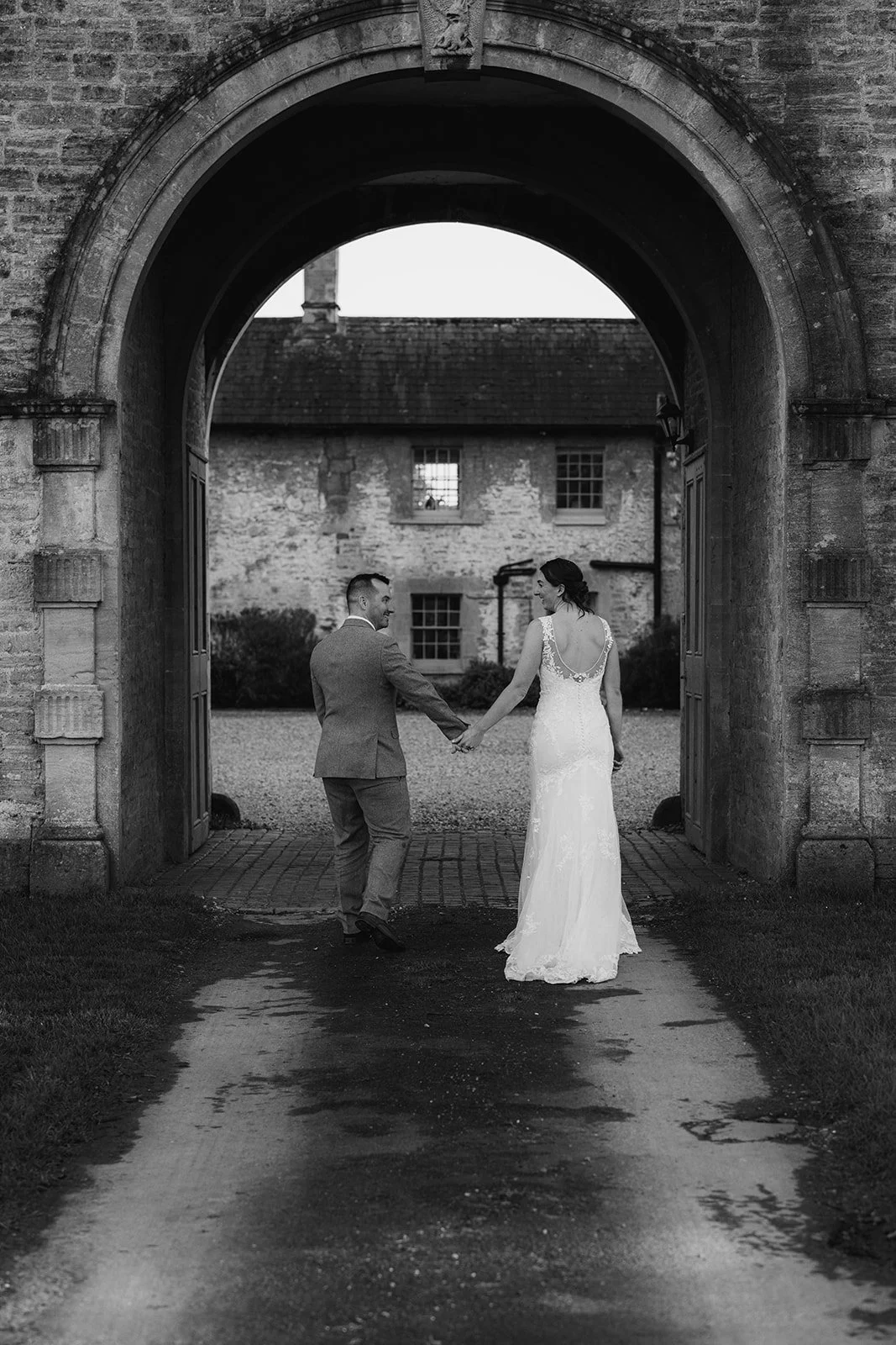 Anna and Chris walk hand in hand beneath the stone archway at Elmhay Park in Somerset, UK, captured in black and white as they look back at one another. A timeless, elegant portrait from Anna & Chris’s wedding day, full of romance and calm.