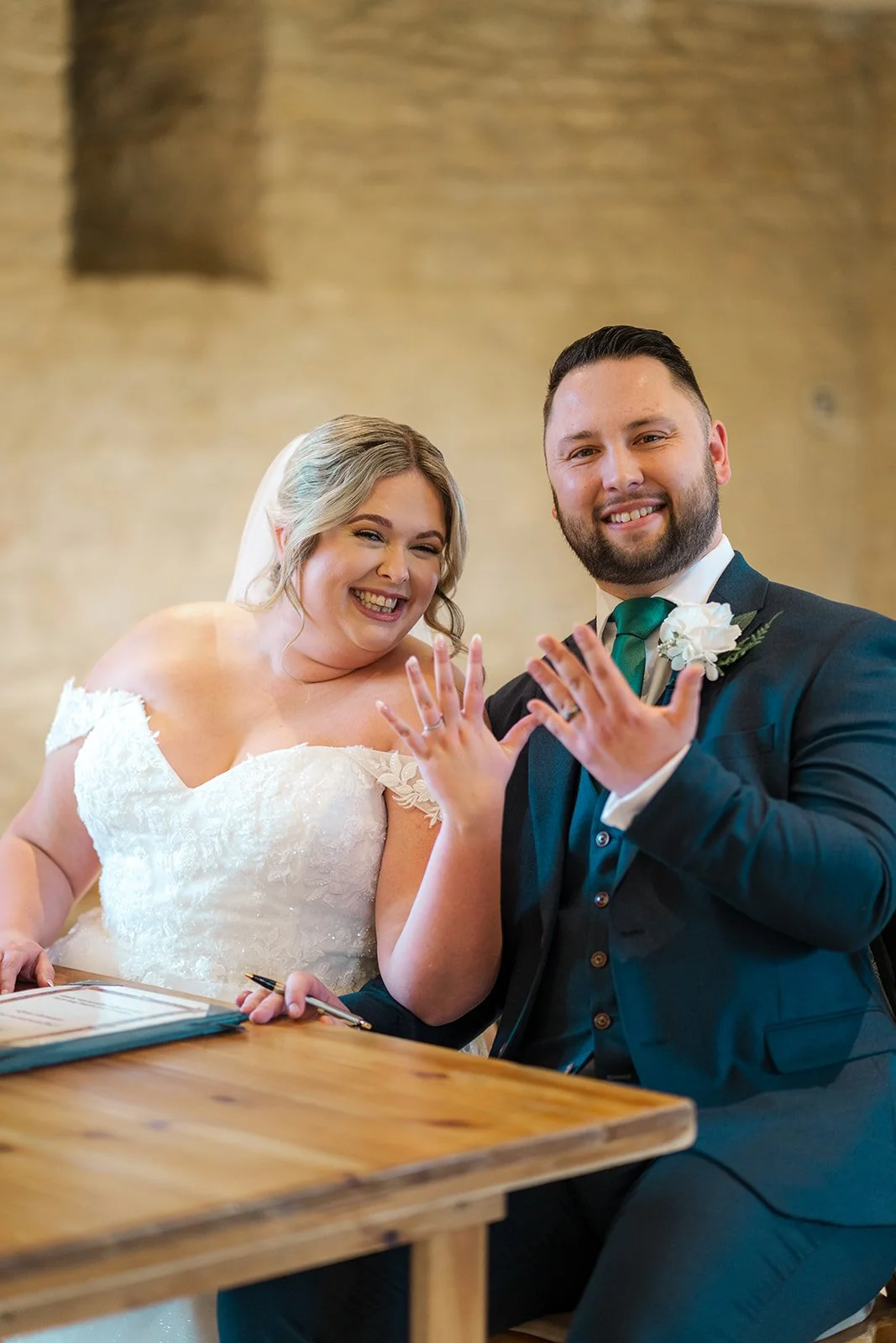 Siobhan & Nicholas sit at the signing table at Kingscote Barn in the Cotswolds, UK, smiling and holding up their wedding rings after the ceremony, a joyful rustic barn wedding moment with the marriage certificate and wooden table in view.