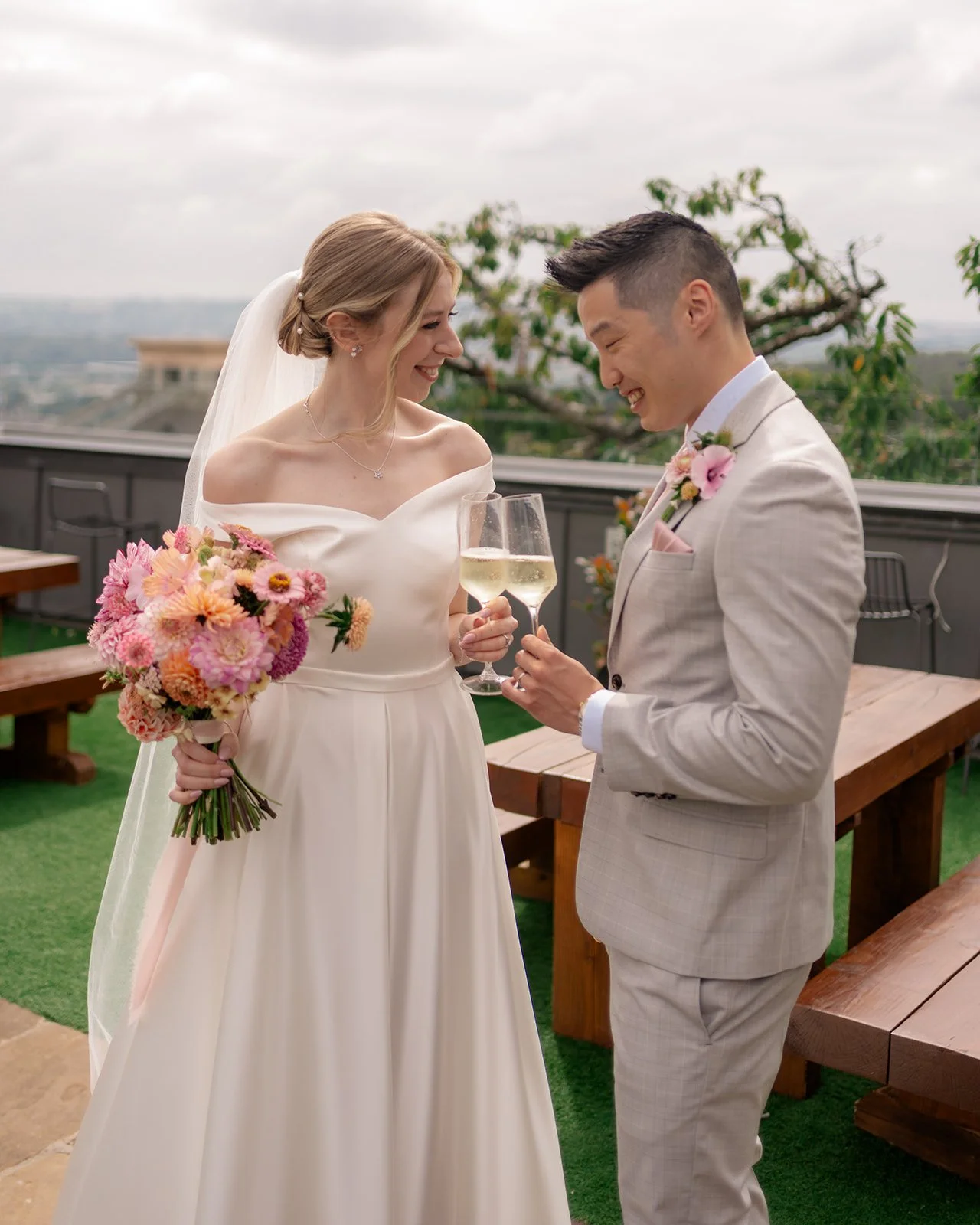 Brian & Josie sharing drinks on the terrace at Clifton Observatory in Somerset, with wedding flowers and views behind them, photographed by Just James Weddings for relaxed couple portraits.