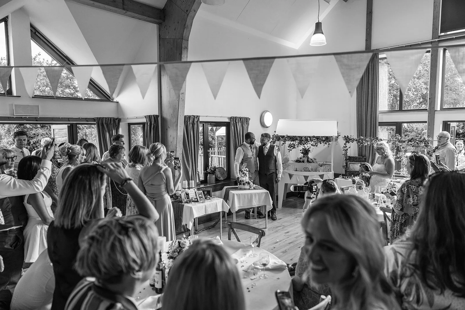 Black-and-white candid of Aaron & Jack surrounded by dancing guests during their lively reception at Rock Village Hall in Worcestershire, photographed by Just James Weddings.