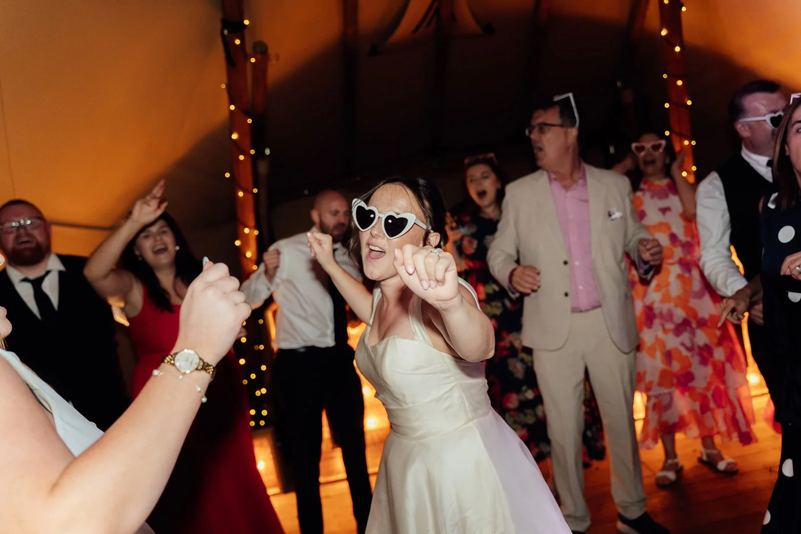 Harriet dances in white heart-shaped sunglasses surrounded by wedding guests during the evening reception at Dovecote Events in Oxfordshire, photographed by Just James Weddings.