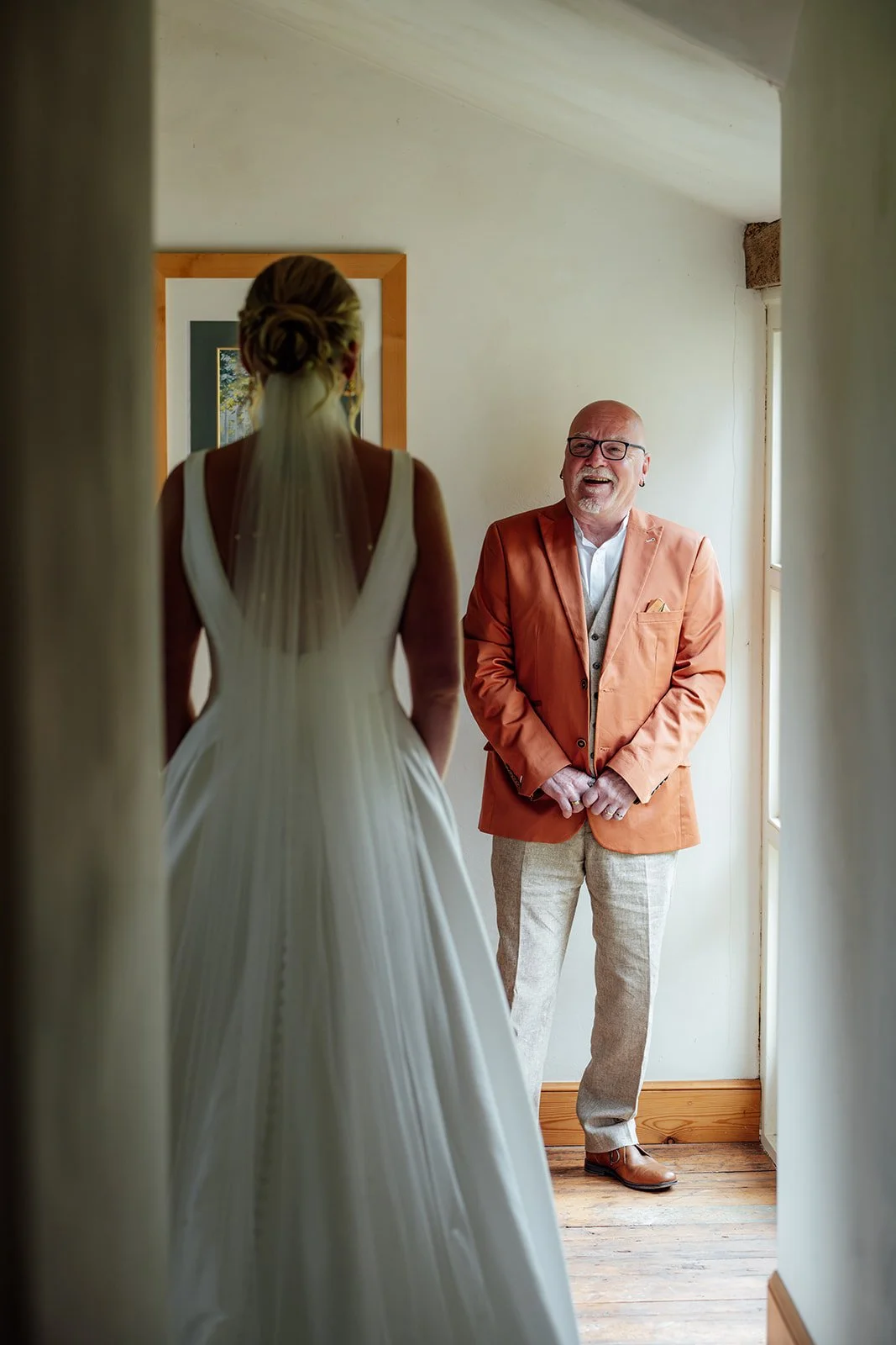 Emotional first look as Hannah reveals her wedding dress to her dad at Grays Barn in Devon, UK, with him smiling in an orange jacket while she stands in her gown and veil in the doorway, photographed naturally by Just James Weddings.