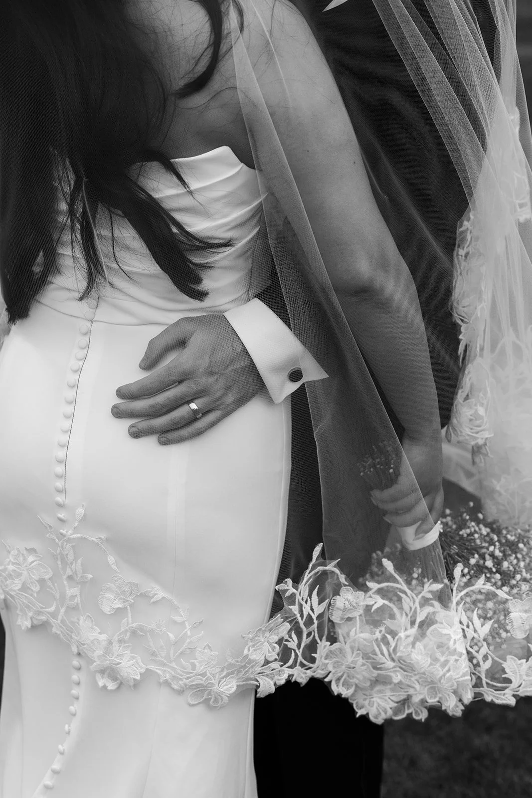 Black and white close-up of Nisha and Jack beneath her lace-edged veil at Batch Country House in Somerset, UK, showing Jack’s hand around her waist, his wedding ring, her dress buttons and delicate bridal details together by Just James Weddings.
