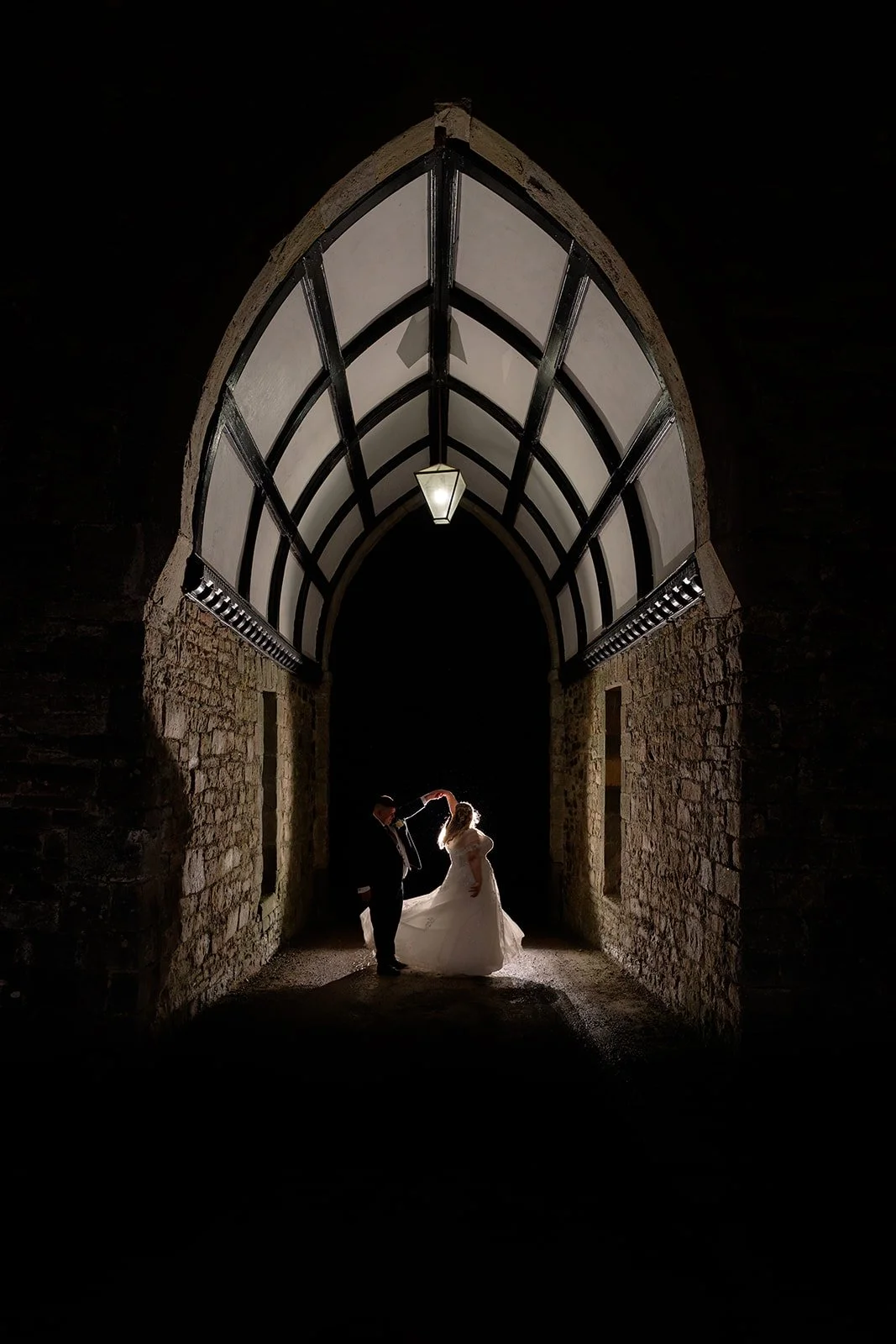 Becky & Rob share a romantic evening portrait beneath the illuminated stone archway at Clearwell Castle in Gloucestershire, UK. Dramatic backlit wedding portrait of the couple dancing under the historic castle arch.