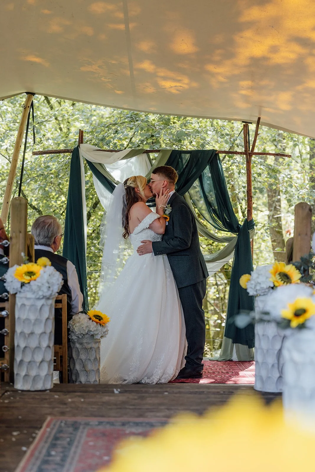 Lucy and Matthew sharing their first kiss beneath the woodland ceremony backdrop at Curated Canvas at Noah’s Pond in Somerset, photographed by Just James Weddings.