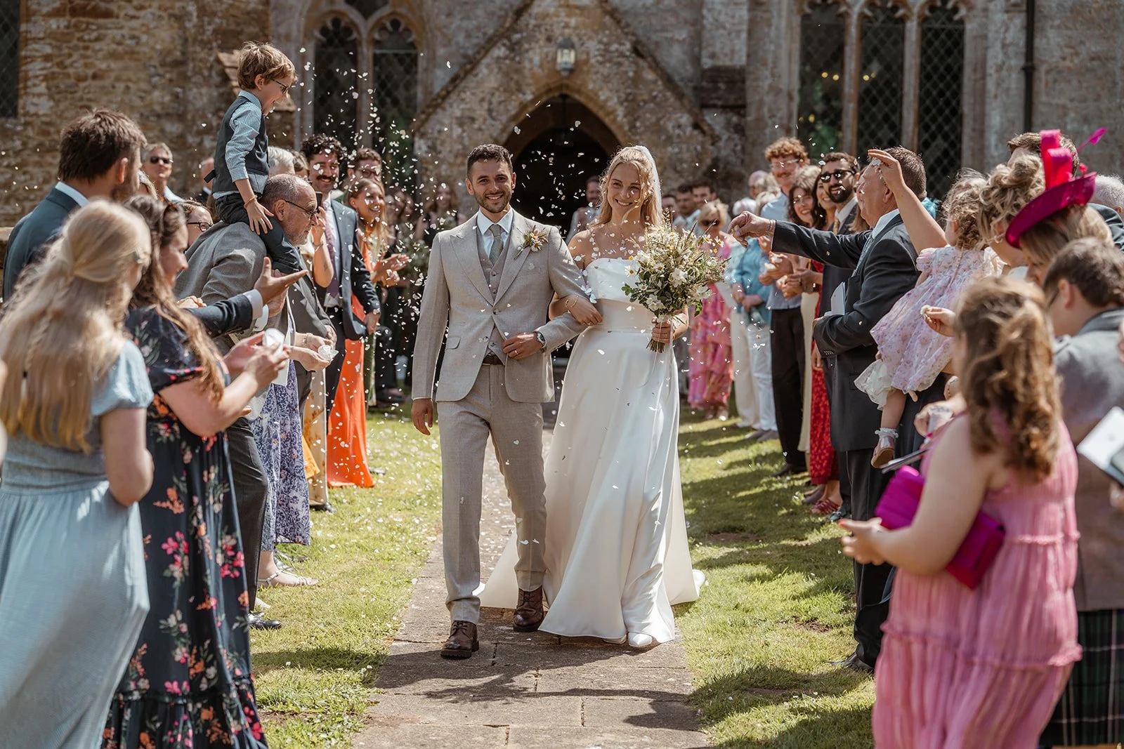 Louise & Christian walking through confetti after their ceremony at The Cider Barn in Somerset, with guests cheering outside the church, photographed by Just James Weddings in a joyful documentary wedding style.
