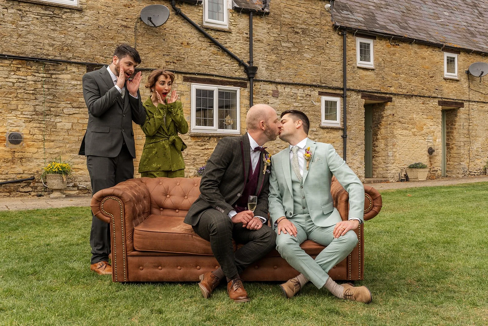 Sam & Gavin kissing on a vintage sofa at The Great Barn at Aynho in the Cotswolds, with guests reacting behind them, photographed by Just James Weddings.