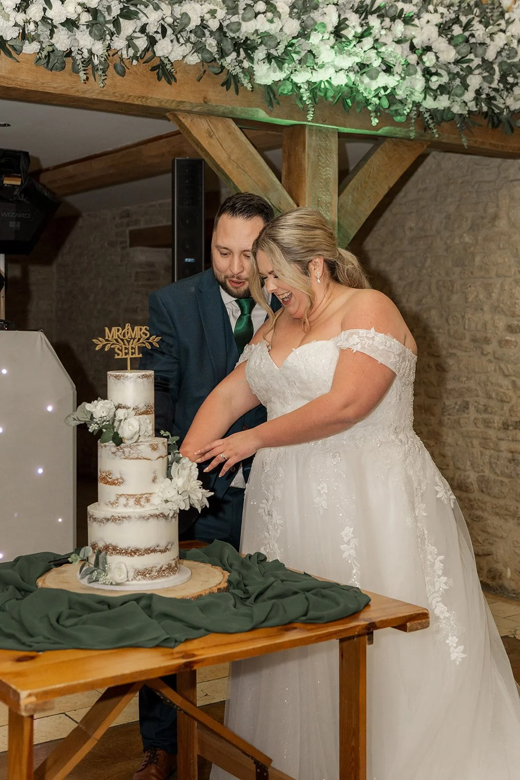 Siobhan & Nicholas cut their wedding cake at Kingscote Barn in the Cotswolds, UK, smiling beside a white tiered cake with floral details, a joyful rustic barn wedding reception moment beneath greenery, wooden beams and warm evening light inside.