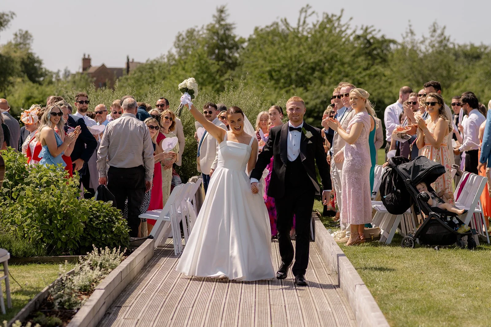 Lydia & Kieran walking hand in hand down the aisle after their outdoor ceremony at Hencote Estate in Shropshire, UK, as guests clap around them in the sunshine, with garden borders, white chairs and countryside greenery behind them in summer.