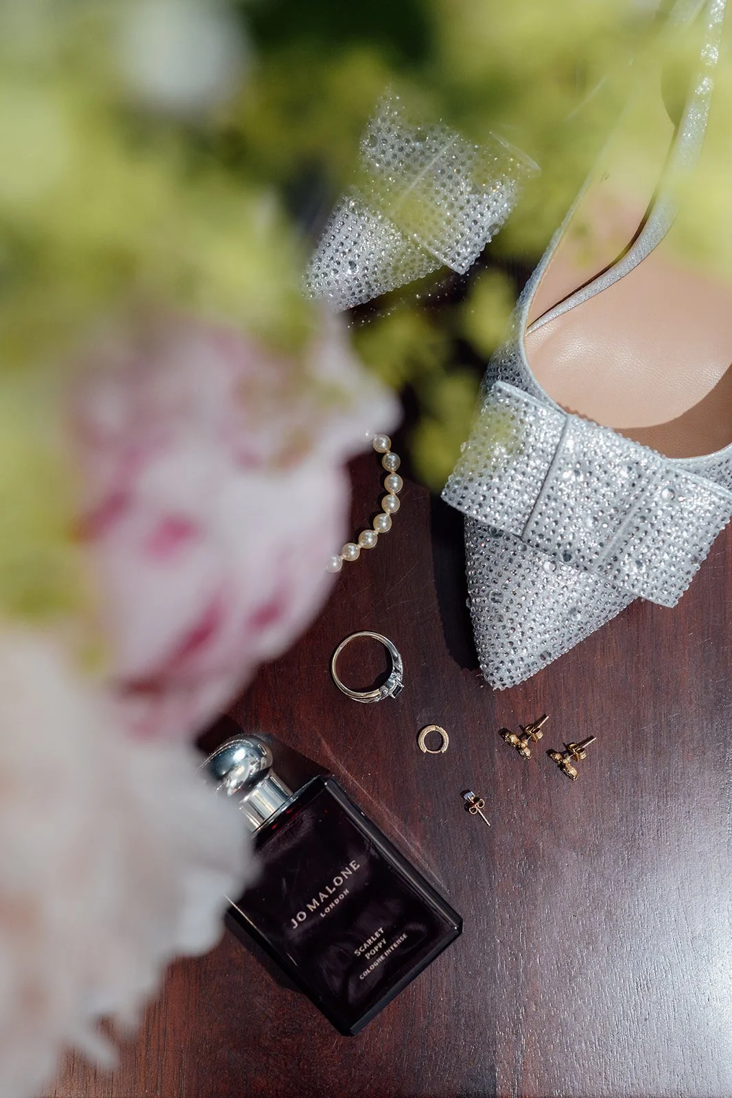 Charlotte & Jack wedding detail photo at Bishop’s Palace in Somerset, UK, showing sparkling bow heels, rings, pearl bracelet, earrings and Jo Malone perfume styled on dark wood with soft flowers in the foreground during bridal preparations.