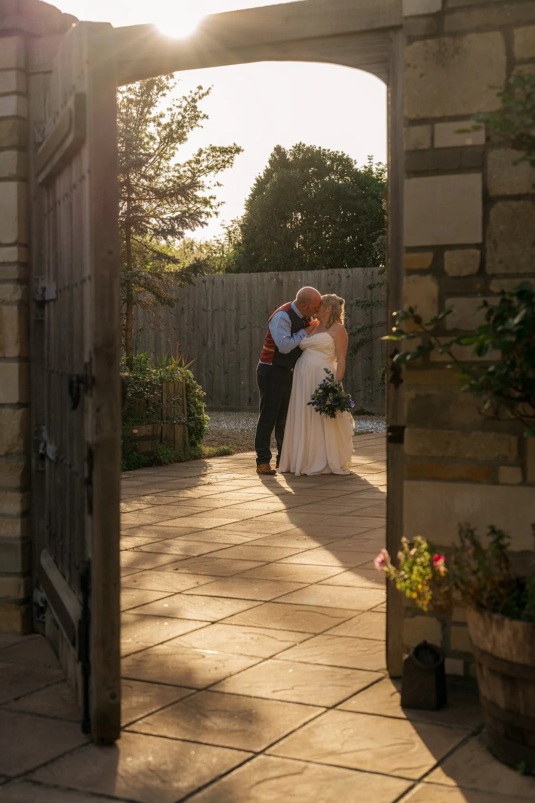 Debbie and Nigel share a golden hour kiss through the open gates at Yarlington Barn in Somerset, with warm evening sunlight, rustic stonework and a quiet courtyard.