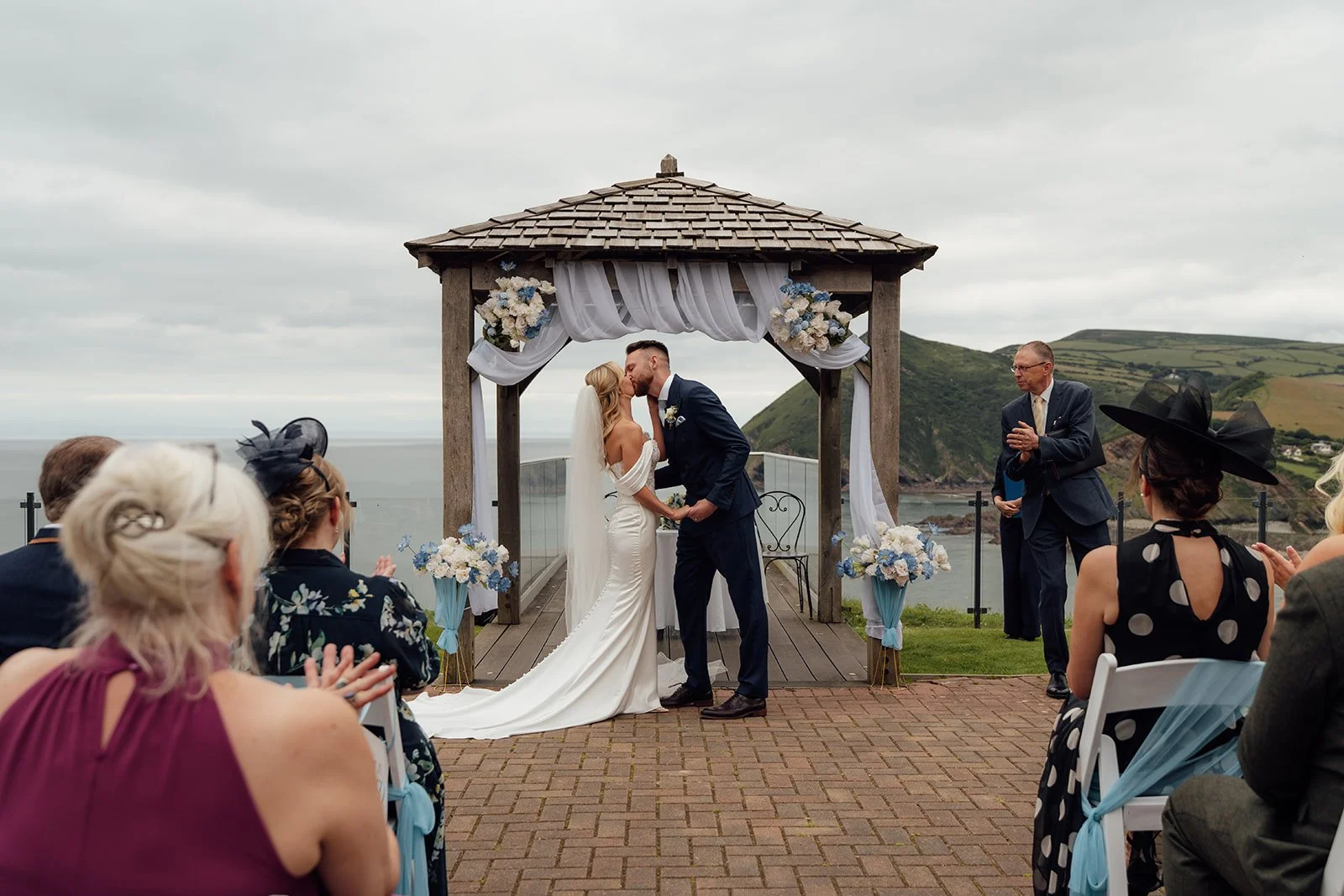 Amy & Connor share their first kiss beneath the flower-decorated outdoor ceremony gazebo as guests applaud at Sandy Cove Hotel in North Devon, UK.