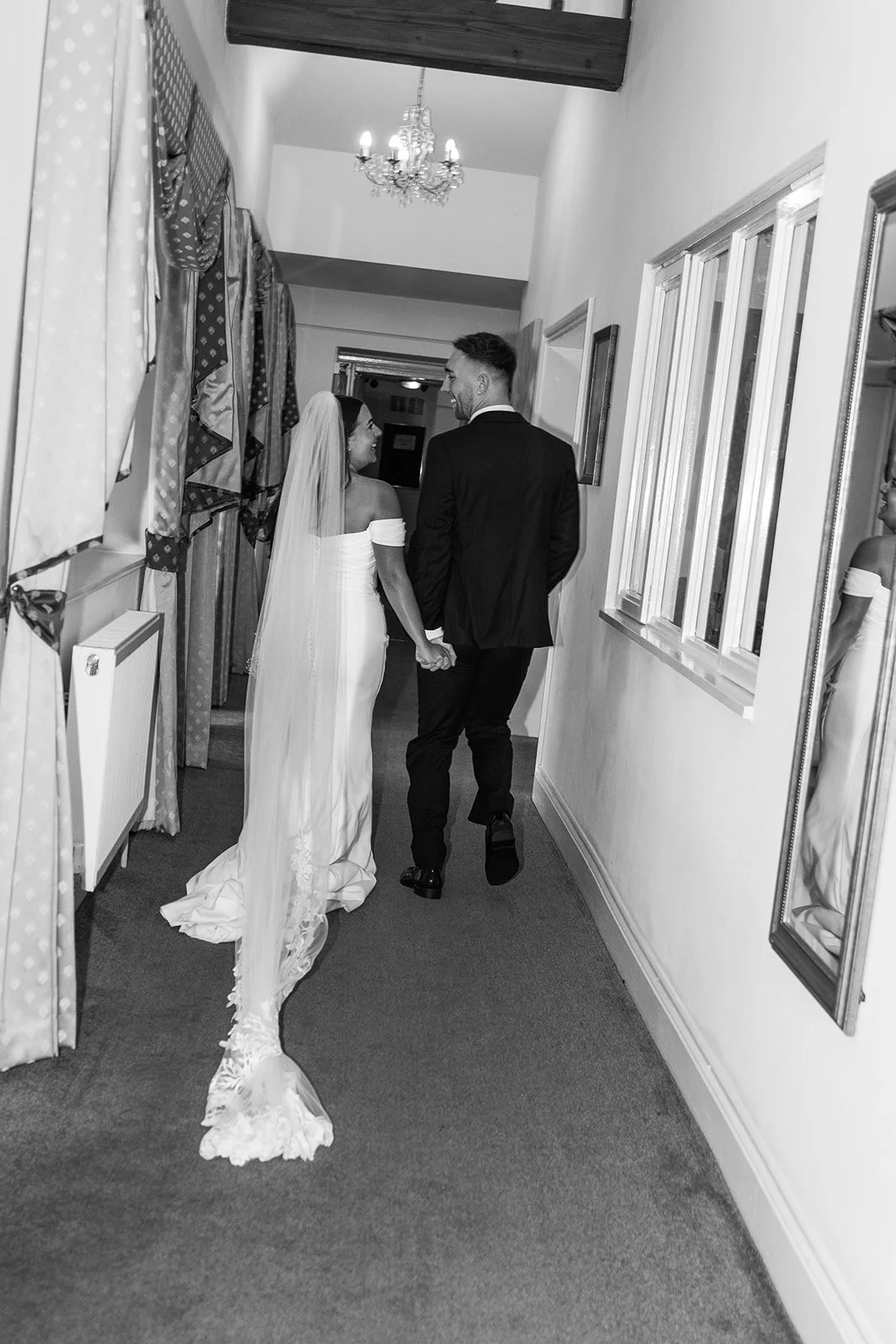 Black and white photo of Nisha and Jack walking hand in hand through Batch Country House in Somerset, UK, with Nisha’s long veil trailing along the hallway as the couple look back at one another, photographed by Just James Weddings.