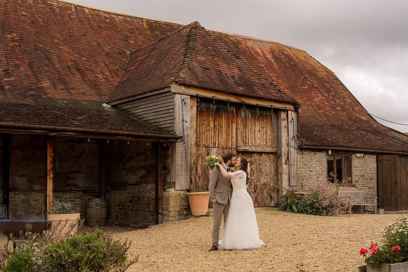 Maisie & Andrew kissing outside the rustic barn at Stockbridge Farm Barn in Dorset, photographed by Just James Weddings for relaxed autumn wedding venue portraits.