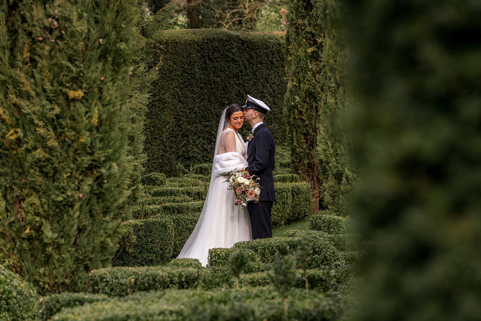 Megan & Luca share a quiet embrace in the garden maze at The Great Barn Devon, framed by tall hedges and neat box borders. Megan rests her head close as Luca leans in for a tender kiss during their Devon wedding portraits.