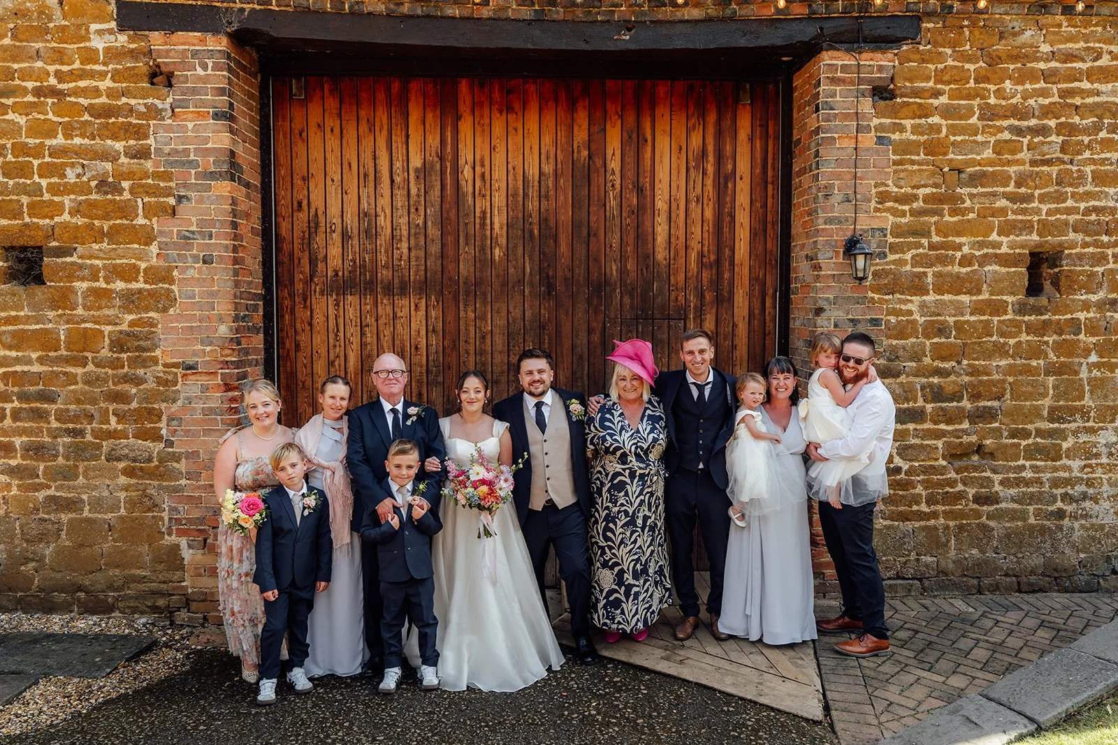 Harriet & Simon pose with family members and children in front of the large wooden barn doors at Dovecote Events in Oxfordshire, photographed by Just James Weddings.